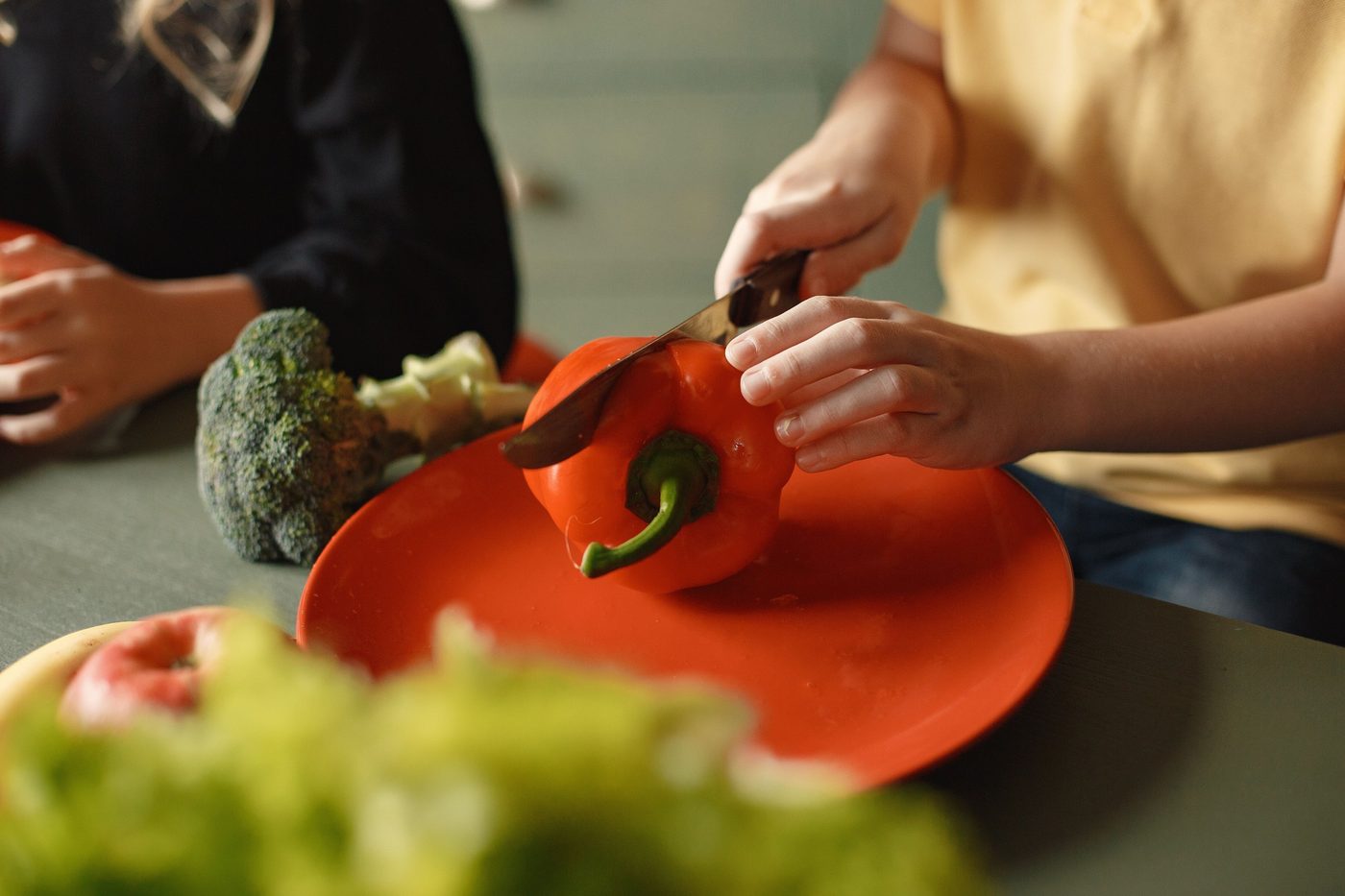 Kinderhände schneiden eine rote Paprika, daneben Brokkoli